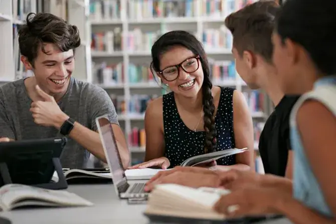 A group of teenagers smiles sitting in a library, surrounded by book and computers on the table.