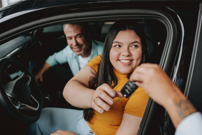 A young woman is seated in the driver’s seat of a car. She smiles as someone hands her the keys to her new car.