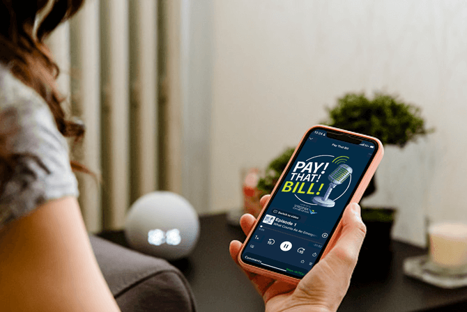 Person holding a smartphone displaying a "Pay That Bill" podcast on screen, seated at a table in a cozy room.