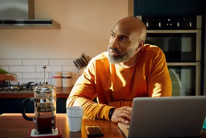 Man standing at kitchen counter with coffee cup and cell phone using laptop and looking to side thinking.