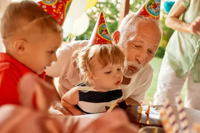 Family celebrating a young child's birthday party outdoors, with an older adult helping the child blow out candles on a cake. Several people, including a child in a party hat, are gathered around the table decorated with party items.