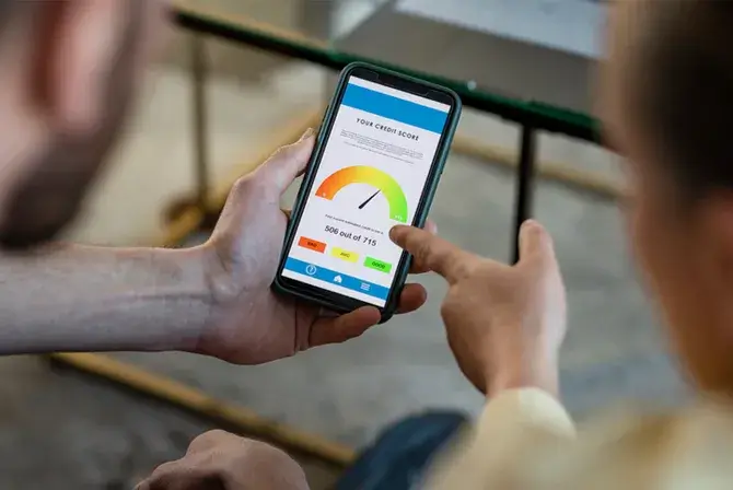A person holds a phone with a credit score graphic displayed on the screen. Their profile is out of focus in the foreground and another person whose profile is also out of focus in the foreground points to the screen as if discussing the score.