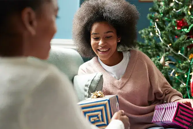 A woman smiles as she receives a wrapped gift from another person who is out of focus in the foreground. A Christmas tree can be seen in the background.