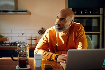 Man standing at kitchen counter with coffee cup and cell phone using laptop and looking to side thinking.