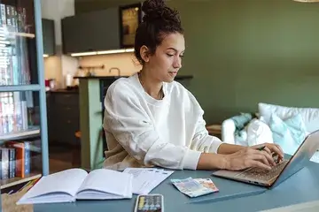 Young woman seated at a table working on her laptop. Money, books and a calculator are visible in the foreground and indicate she could be working on a budget.