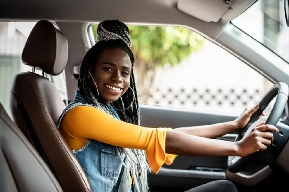 Young woman sitting in the driver’s seat of a car with both hands on the steering wheel, smiling at the camera.