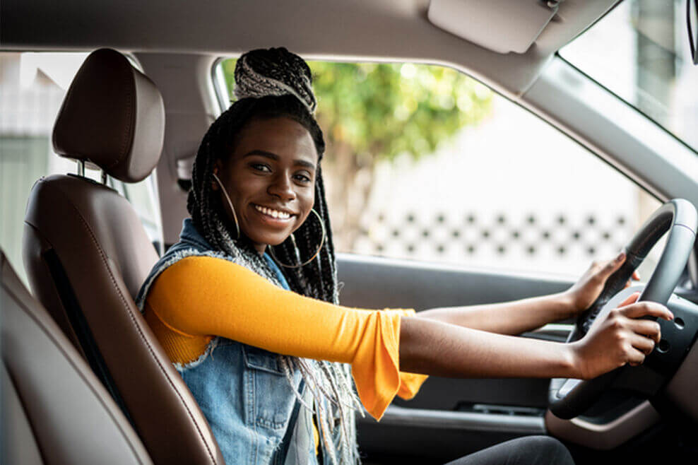 Young woman sitting in the driver’s seat of a car with both hands on the steering wheel, smiling at the camera.