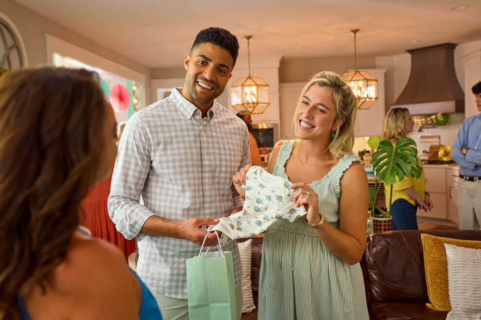 A couple holds up a baby onesie in excitement at a baby shower.