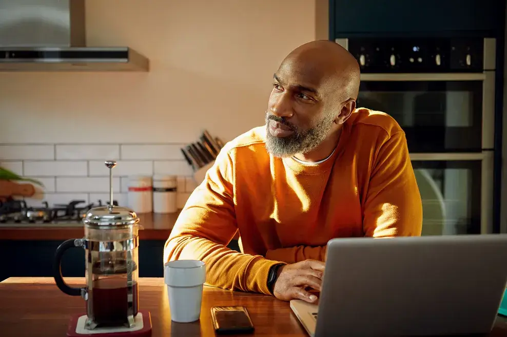 Man standing at kitchen counter with coffee cup and cell phone using laptop and looking to side thinking.