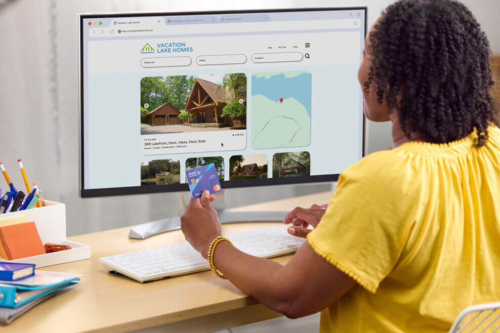 Person holding a VACU credit card while browsing a vacation home rental website on a computer screen in an office setting.