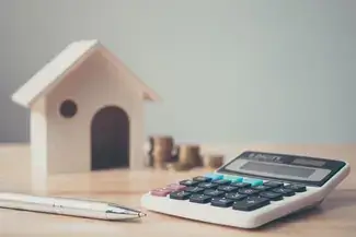 calculator and pen with a mini house and coins in the background
