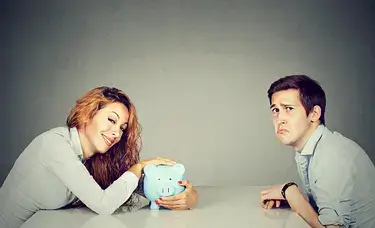 A woman smugly holding a piggy bank across the table from a sad looking man.