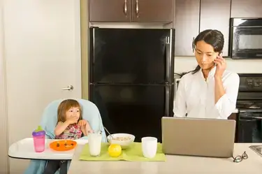 A mom on a laptop and her young daughter in a high chair in a kitchen.