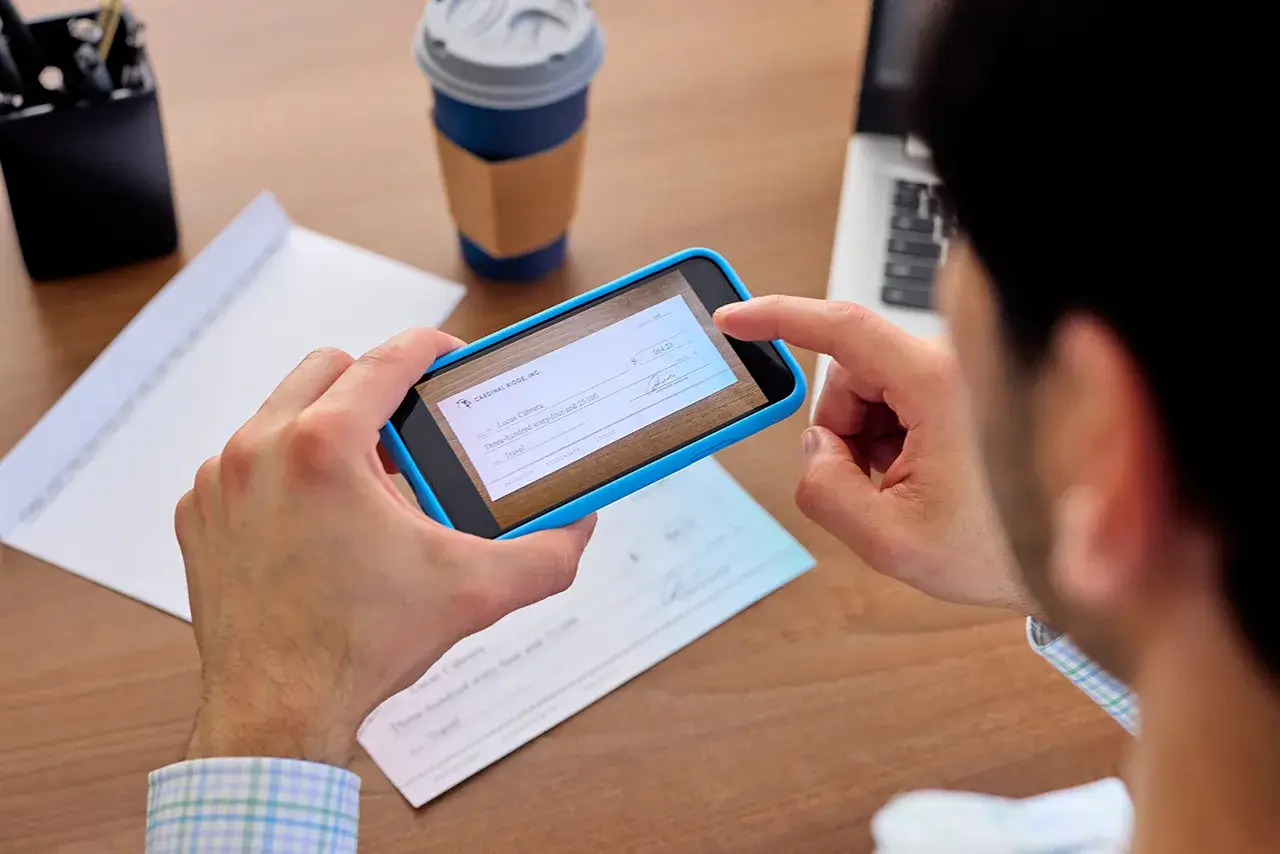 Individual holding phone and taking a picture of a check on a desk, with a coffee and computer in the background