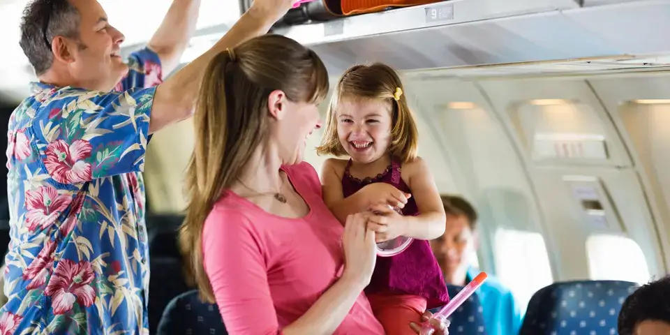 Woman and young child smiling while boarding a plane, while man behind them loads luggage into overhead storage.
