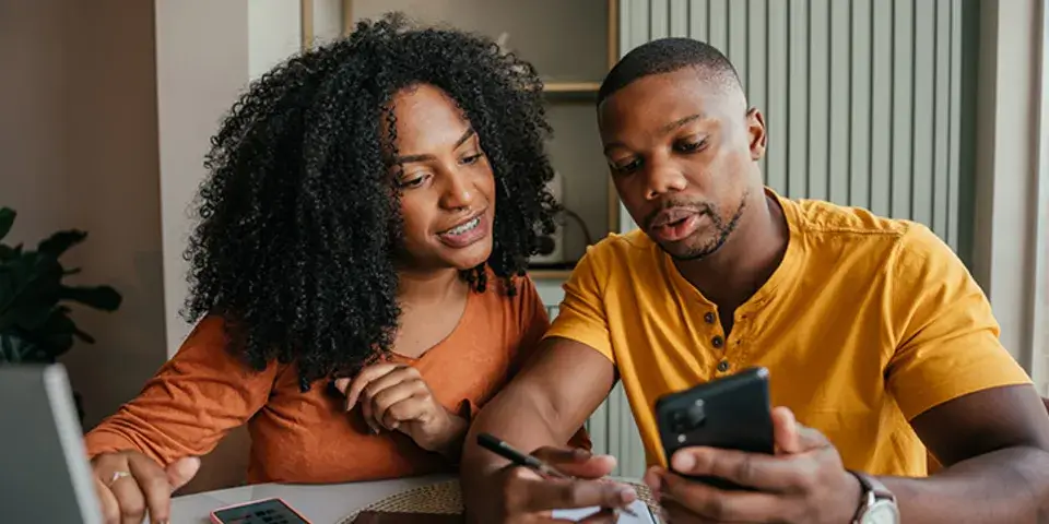 A woman and a man sit at a table while writing on paper and looking at a mobile device.