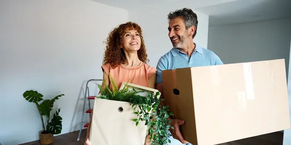 Man and woman standing inside house, holding boxes and smiling