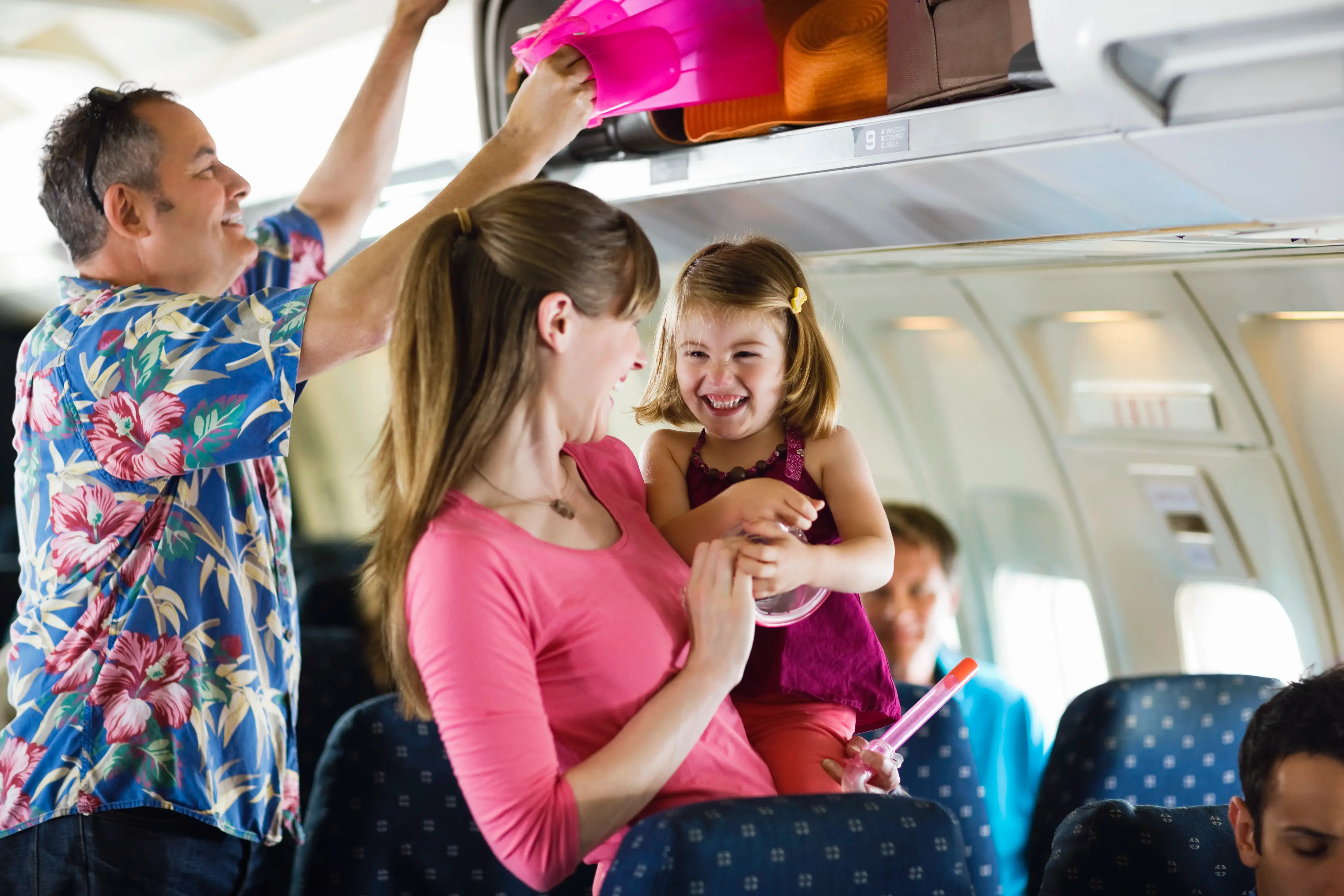 Woman and young child smiling while boarding a plane, while man behind them loads luggage into overhead storage.