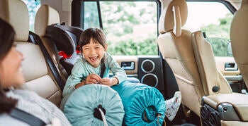 Smiling child seated in a car’s back seat next to a car seat and travel pillows, laughing during a road trip