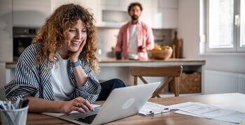 Women working at kitchen table on a laptop, surrounded by papers. Man stands at counter in the background.