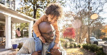 Man carrying a smiling child on his shoulders outside their home.